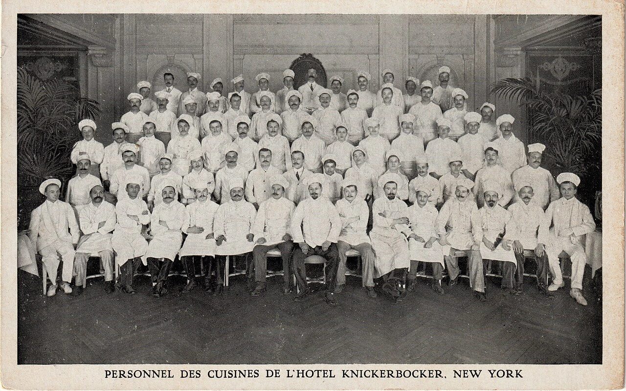 A large kitchen staff posed in rows in a hotel dining room, nearly all in white jackets and toques, with a few men in dark suits