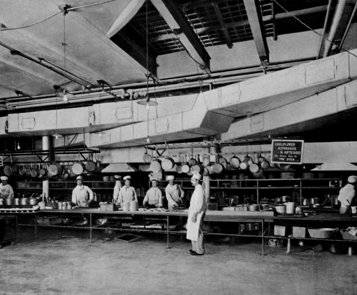 A large hotel kitchen: cooks in white jackets and toques stand along a long work table under racks of hanging pans, with a sign reading Cauliflower, Asparagus and Artichoke