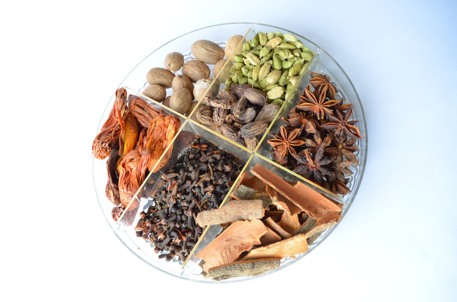A round glass dish divided into sections of whole spices: nutmeg, green and black cardamom, star anise, mace, cloves and pieces of cinnamon bark